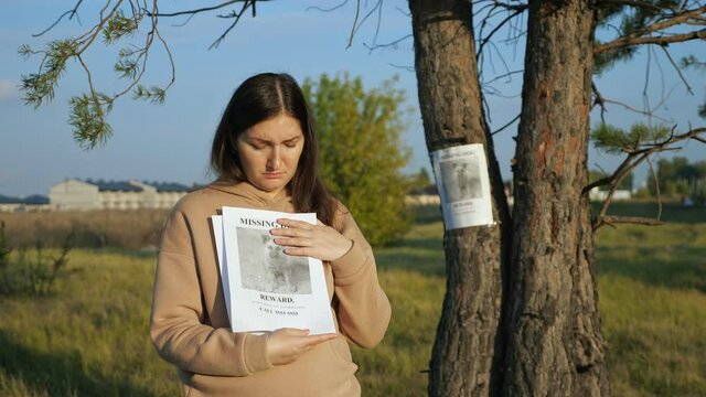 woman with flyers stack stands near poster report of missing dog with photo stuck on tree trunk closeup
