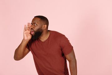 Funny young African-American man in brown t-shirt talks covering mouth by palm on light pink background in studio close view
