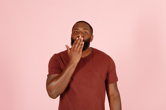 Handsome young African-American man model in casual brown t-shirt covers his mouth with his hand on light pink background in studio
