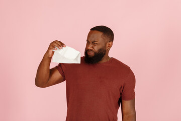 Disgusted African-American man with nasal cold looks at dirty wet tissue standing on light pink...
