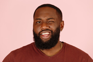 Obraz premium Attractive bearded African-American man model in brown t-shirt winks standing on light pink background in studio close view