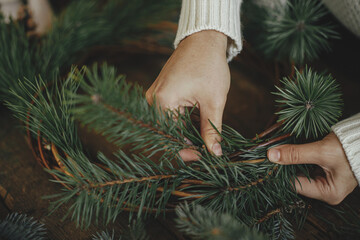 Woman hands holding pine branches and arranging christmas wreath on rustic wooden background. Winter holiday preparations. Festive workshop. Making rustic christmas wreath close up