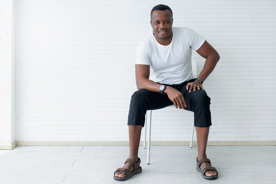 African American Man Wearing A White T-shirt Sits In A Chair And Looks At The Camera On A White Background., Happy Confident Black Guy Posing For Full Body