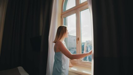 Young dreamy woman in sleepwear opening curtains of window in cozy bedroom. Elegant caucasian girl in apartment enjoying cityscape views and sunshine.