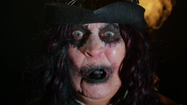 Sinister elderly woman with horrible scary Halloween witch makeup in costume making faces, looking ominous at camera, trying to scare. Portrait of vampire granny isolated against black background