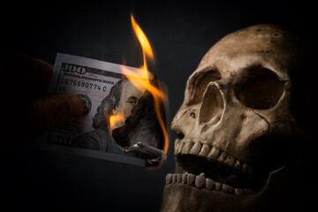 Fingers hold a burning USD 100 banknote next to a dummy human skull on a black background