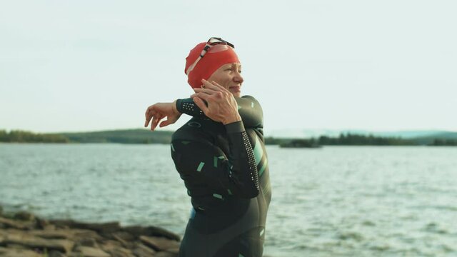 Female triathlete in swimsuit standing on lakeshore and doing stretching exercises while preparing for swimming workout