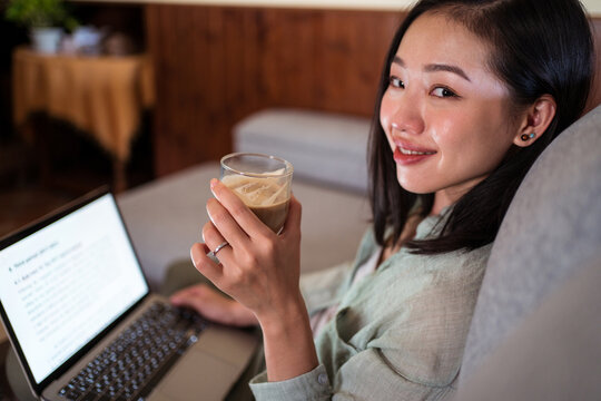 Asian freelancer typing on laptop on couch while drinking latte