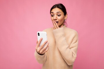 Photo shot of surprised amazed shocked attractive positive good looking young woman wearing casual stylish outfit poising isolated on background with empty space holding in hand and using mobile phone