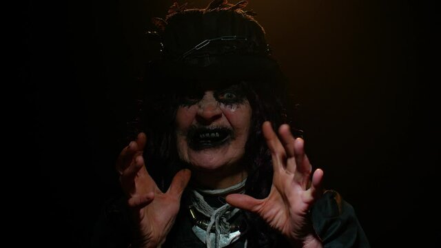 Sinister elderly woman with horrible scary Halloween witch makeup in costume making faces, looking ominous at camera, trying to scare. Portrait of vampire granny isolated against black background