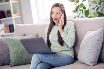 Photo of cheerful millennial brunette lady sit on sofa with laptop talk telephone wear blue shirt at home alone