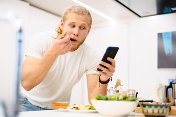 Young white man using cellphone while having lunch in kitchen