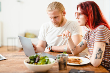 Young white couple using laptop and having lunch while sitting at table