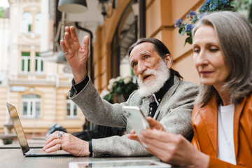 White senior couple gesturing and using gadgets while sitting in cafe