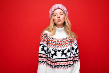 Photo of pretty positive cute joyful young blonde woman isolated over red background wall wearing winter sweater and pink hat giving kiss