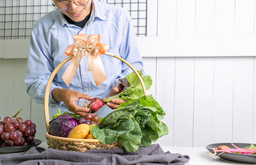 A smiling Asian man in a blue shirt holds a basket of fresh vegetables, kale, potatoes, purple cabbage, and grapes. Gifts for health Concept