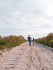 Happy father and daughter running jumping having fun on empty autumn sea beach. Dad and child walking on white sand road with reeds blue sky background. Lifestyle real people. Happiness travel concept