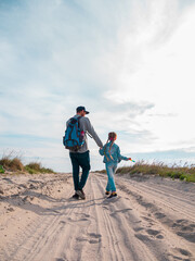 Happy father and daughter running jumping having fun on empty autumn sea beach. Dad and child walking on white sand road with reeds blue sky background. Lifestyle real people. Happiness travel concept
