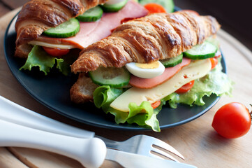 Croissant sandwich with ham, cheese, fresh salad, tomatoes, eggs and cucumber on wooden background. Breakfast with fresh croissant sandwich