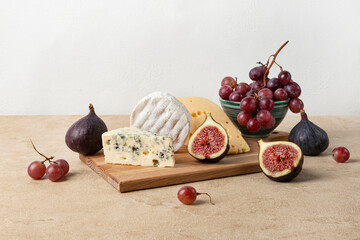 Camembert cheese, maasdam cheese and cheese with blue mold lie on a wooden board surrounded by figs and grapes. Horizontal orientation, beige textured background.