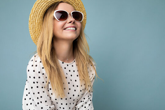 Closeup shot of fascinating smiling positive young blonde woman wearing summer dress straw hat and stylish sunglasses isolated over blue background wall looking to the side
