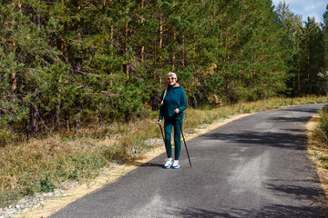 happy sixty-year-old woman in sportswear and sneakers walks along the track with poles for Nordic walking