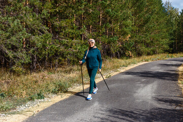 happy sixty-year-old woman in sportswear and sneakers walks along the track with poles for Nordic walking