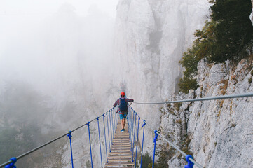 A young man in mountaineering equipment walks uphill on a suspension bridge in the fog. Suspension bridge on Mount Ai-Petri, Yalta, Crimea. The concept of travel, active lifestyle, tourism. - obrazy, fototapety, plakaty