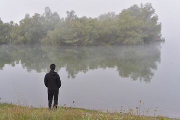 Fototapeta premium man stands early in the morning on the shore of the lake in the fog