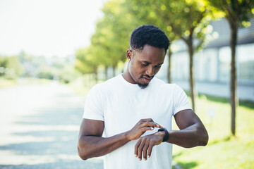Young african american man, on a morning jog in a white t-shirt in the park, switches sports program in smart watch fitness bracelet engaged in fitness active lifestyle