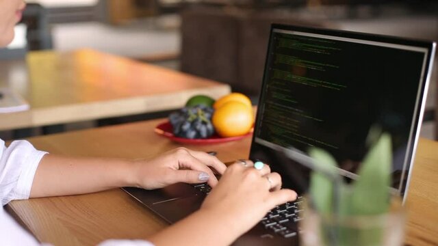 Back view over the shoulder shot of developer programmer female with laptop. Program code and script data on the screen. Young freelancer woman working on project in cafe.