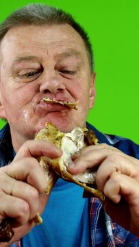 Hungry senior adult caucasian male with pleasure eating fried chicken leg, gnawing teeth into meat and wiping his mouth with his hand.