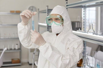 Scientist working in modern science laboratory. Portrait of serious young male researcher in white PPE suit, gloves, face mask and protective goggles looking at sample glass tube he's holding in hands