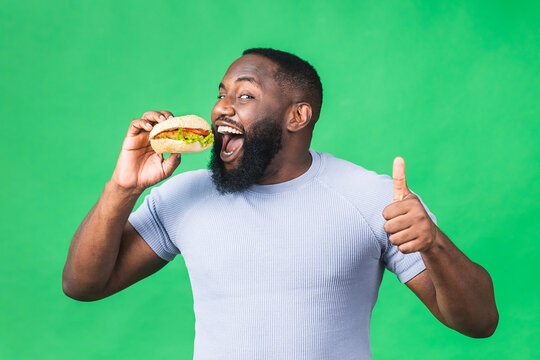 Hungry Young African American Black Man Eating Hamburger Isolated Over Green Background. Diet Concept.