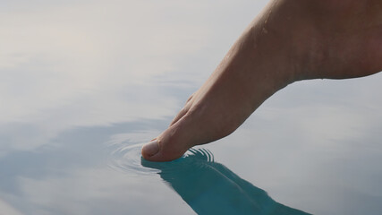 A close-up of a person's foot gently touching the surface of the water is purple-green. A person checks the temperature of the sea with his foot, putting his toe under the water, during . Slow-motion.