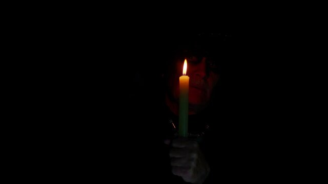 Frightening senior man with Halloween witcher makeup looking at candle, conjure, hex, wiz. Horror theme. Sinister sorcerer guy on studio black background. Voodoo magical creepy rituals with candle
