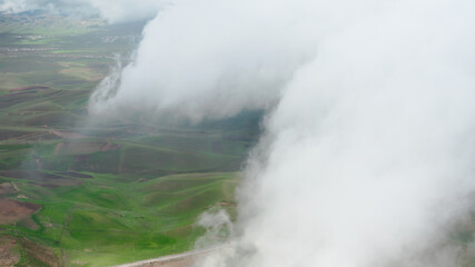 A sky shot shot through rain clouds with a view of green and brown earth fields. A bird's-eye view above the clouds with a view of the road and a rural valley on a sunny day. Aerial copter frame.