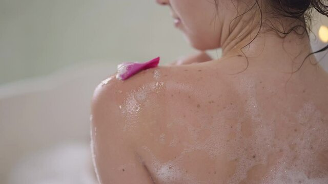 Close-up shoulder of slim young unrecognizable Caucasian woman washing skin with flower petal in slow motion. Sensual smiling gorgeous confident lady taking bath at home indoors. pampering and hygiene