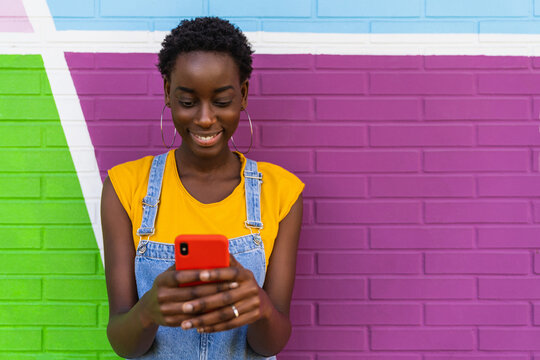 Smiling black woman texting on smartphone near colorful wall