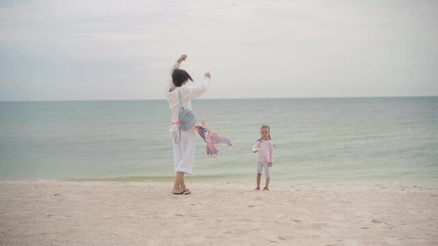 Mother with Little Daughter Dressed White Clothes Playing Kite Sea Beach Happy Caucasian Family With One Child Have Fun Summer Vacation Sea Shore. Active Leisure Positive Emotions Happy Childhood 