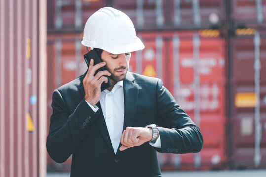 Busy business man, engineer manager wearing white hardhat, looking at his wristwatch, checking the time while standing in front of cargo containers. Waste time reduction engineering site concept.