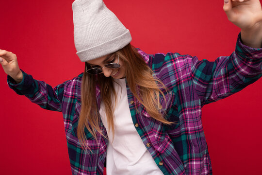Fantastic photo shot of beautiful young dark blonde woman isolated over red background wearing hipster purple casual shirt grey hat and stylish sunglasses looking down and dancing