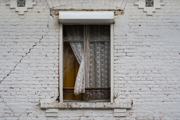 Fototapeta na wymiar Fenster mit eingeklemmter Gardine an einem verlassenen Haus in Nordfrankreich. Weiss gestrichene Ziegelmauer in fortgeschrittenem Stadium der Verwitterung