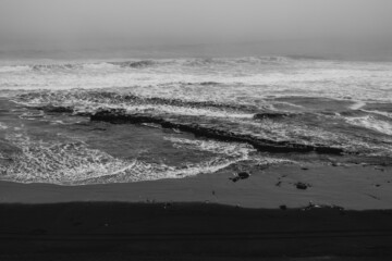 Panoramic view of the shore, the pacific ocean with waves, the horizon with cloudy sky and beach with rocks (in black and white)