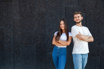 Emotions, gesture, expression and people concept - Happy Caucasian man and woman in white T-shirts and jeans standing next to each other and crossing arms on a black background with copy space on left