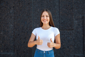 Expression, gesture, people concept - Happy Caucasian girl showing thumbs up, looking at the camera and dressed in a white T-shirt and jeans outdoors on a black background with copy space