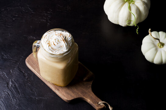 Spicy pumpkin latte in a glass with cream and cinnamon. Cozy warm autumn drink concept with pumpkins on black background in dark key, selective background