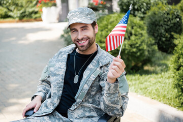 disabled veteran in military uniform holding small usa flag and smiling on city street - obrazy, fototapety, plakaty