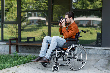 Obraz premium young disabled man in wheelchair taking photo on vintage camera near building with glass facade