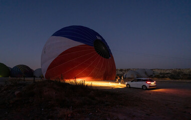 A car parked in front of a hot air balloon with headlights on during night, preparation of a flight in Goreme national park in Cappadocia, Turkey